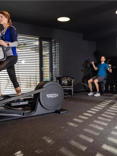Een moderne sportschoolscene met twee personen. Een vrouw traint op een crosstrainer, terwijl een man gewichten heft.