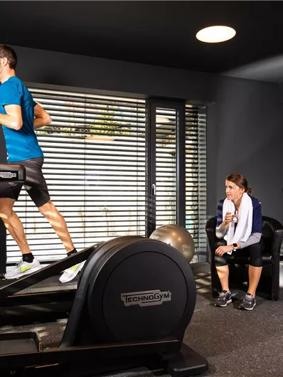 Een man loopt op een loopband in een moderne fitnessruimte. Een vrouw zit ernaast en kijkt toe.