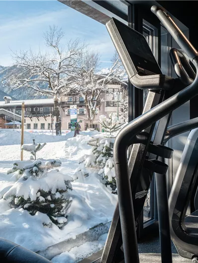A view from a fitness room over a snowy landscape. In the foreground, there is a piece of exercise equipment, and in the background, you can see snow-covered trees and mountains.