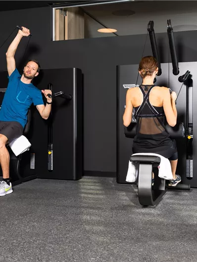 Two people are exercising in a modern gym. One person is using an upper body training machine while the other is training on a stationary bike.