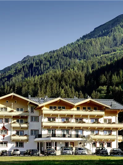 Ein gemütliches Hotel umgeben von grünen Bergen und Wäldern. Die Fassade ist hell und einladend mit Balkonen und Parkplätzen.
