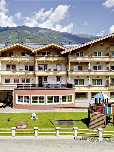 Ein modernes Hotelgebäude umgeben von grünen Wiesen und Bergen. Im Vordergrund befinden sich ein Spielplatz und eine Terrasse zum Entspannen.
