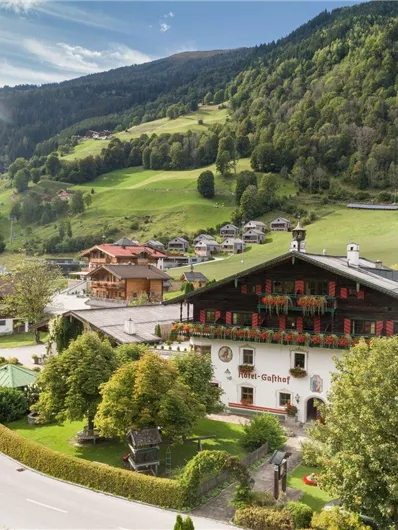 Ein malerisches Alpenpanorama mit grünen Wiesen und Bergen. Im Vordergrund steht ein traditionelles Gebäude, umgeben von Bäumen und einer ruhigen Landschaft.