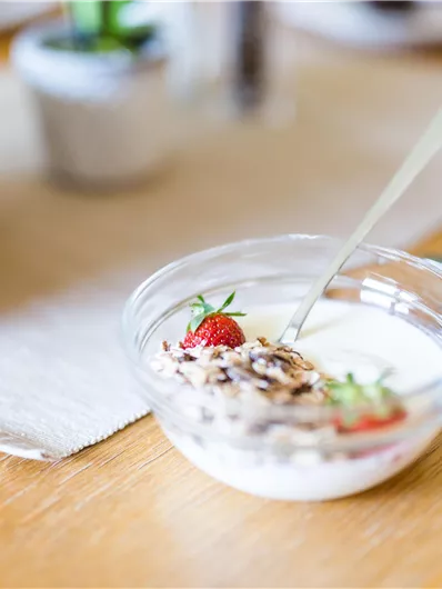 Ein Glas Joghurt mit Haferflocken und frischen Erdbeeren steht auf einem Holztisch. Nebenan befindet sich ein Korb mit Brötchen und ein gedeckter Tisch.