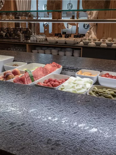 Eine Auswahl frischer Früchte in Schalen auf einer Theke. Die bunten Früchte umfassen Wassermelonen, Pfirsiche, Trauben und Tomaten.