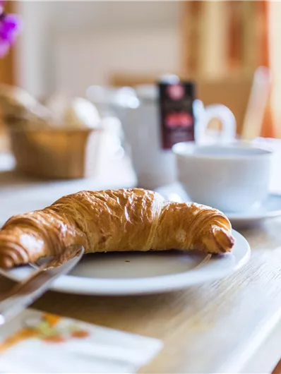 Ein frisch gebackenes Croissant liegt auf einem Teller. Im Hintergrund sind eine Tasse Kaffee und einige Gewürze zu sehen.