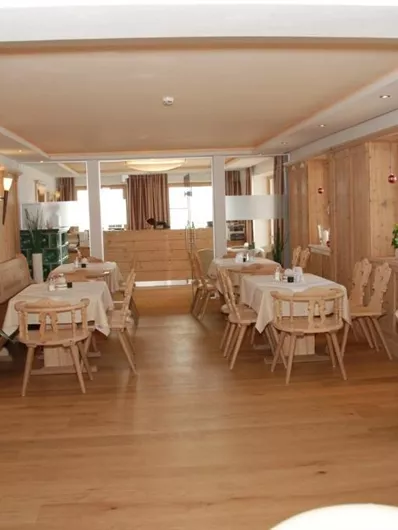 A cozy restaurant with wooden paneling and bright lighting. There are tables and chairs as well as some plants placed in the room.