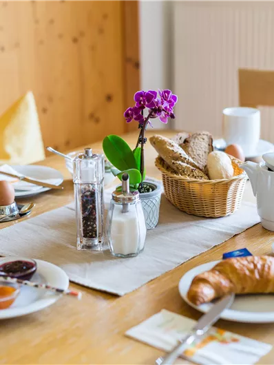 Ein gemütlicher Frühstückstisch mit verschiedenen Brötchen, Croissants und Marmelade. Eine hübsche Orchidee und Getränke runden das Frühstücksbuffet ab.