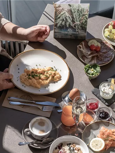 Ein reichhaltiges Frühstücksbuffet mit verschiedenen Speisen, darunter Eier, frisches Obst und Aufschnitt. Eine Person sitzt am Tisch und serviert sich ein Gericht.