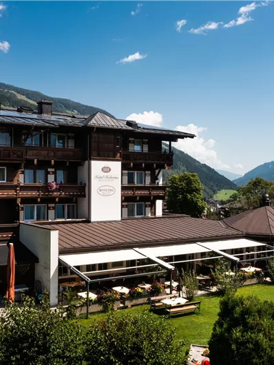 Een charmant hotel in Alpine stijl met een mooie tuin. Op de achtergrond zijn groene heuvels en een blauwe lucht te zien.