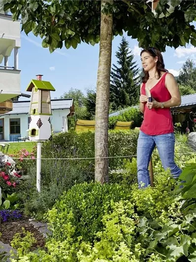 Een vrouw wandelt door een mooie tuin met kleurrijke bloemen en een vogelhuisje. Op de achtergrond ziet men een modern huis en zachte heuvels.