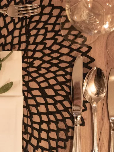 Ein elegant gedeckter Tisch mit Besteck und Gläsern auf einem dekorativen Platzset. Eine Serviette mit einem grünen Zweig verleiht dem Arrangement einen frischen Akzent.