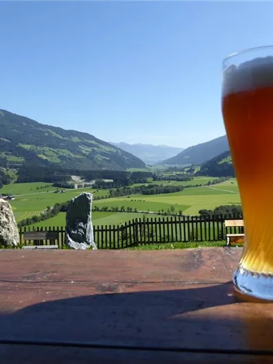 Ein Glas Bier steht auf einem Tisch mit Blick auf das grüne Tal und die Berge im Hintergrund. Der Himmel ist klar und blau, was eine entspannte Atmosphäre schafft.