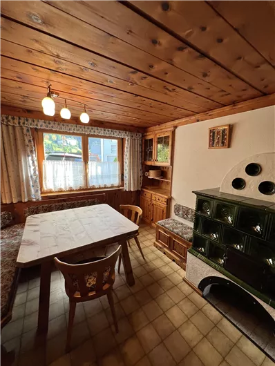 A cozy dining area with wooden ceilings and a large table. In the corner stands a traditional stove and beautiful windows with curtains.