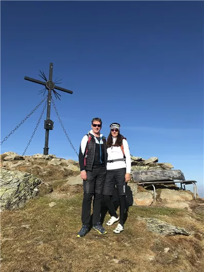 A couple stands on a mountain peak with a cross in the background. The sky is clear and sunny.