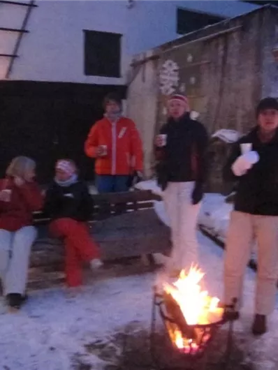 Eine Gruppe von fünf Personen steht und sitzt um ein Lagerfeuer im Schnee. Sie tragen warme Kleidung und scheinen eine gemütliche Zeit zusammen zu haben.