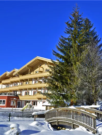Ein charmantes Hotel im Schnee mit einem großen Baum im Vordergrund. Der Himmel ist klar und blau, während die Berge im Hintergrund zu sehen sind.