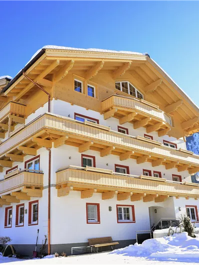 Ein schönes Skihaus im Schnee mit mehreren Balkonen. Umgeben von einer winterlichen Landschaft und blauen Bergen.