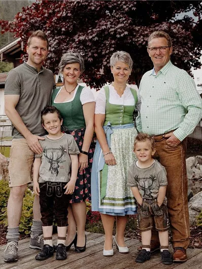 Eine Familie steht in traditioneller Tracht vor einem malerischen Hintergrund. Im Vordergrund sind zwei Kinder und im Hintergrund sind Häuser und eine schöne Gartenlandschaft zu sehen.