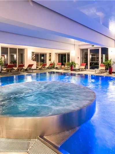 Ein moderner Poolbereich mit einem runden Whirlpool und vielen Liegen. Große Fenster lassen viel Tageslicht herein und bieten einen schönen Ausblick.