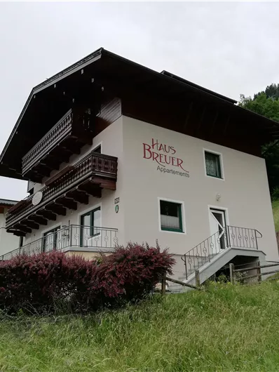 Een modern vakantiehuis met de naam "Haus Breuer" te midden van een groene landschap. De gevel is licht en heeft een overdekt balkon.