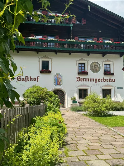 Ein traditionelles Gasthaus mit weißer Fassade und grünem Balkon. Der Weg ist von üppigem Grün und Blumen gesäumt.
