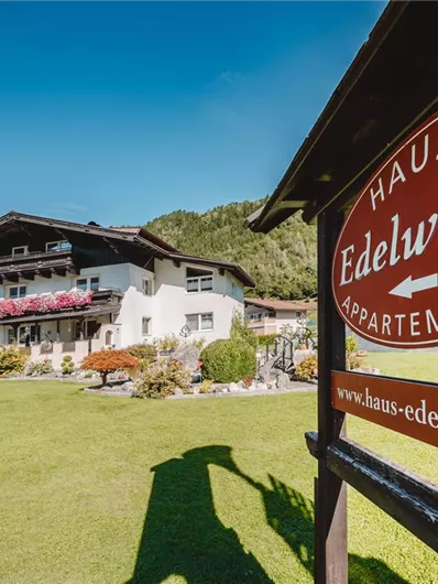 A beautiful chalet with a well-kept garden and colorful flowers. In the foreground, there is a sign that reads "Haus Edelweiss Appartements".