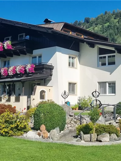 A beautiful house with a balcony and blooming plants. The garden is well-kept and surrounded by mountains.