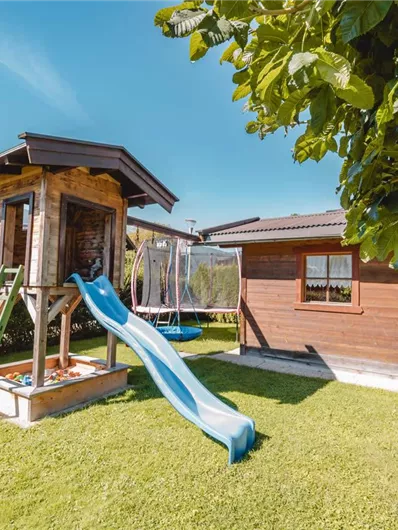 A beautiful garden with a playground, a slide, and a small wooden cabin. In the background, houses and trees are visible.