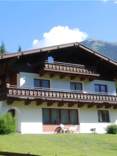 Ein traditionelles Haus in den Alpen mit einem Balkon und viel Grün im Vordergrund. Im Hintergrund sind Wälder und Berge sichtbar.