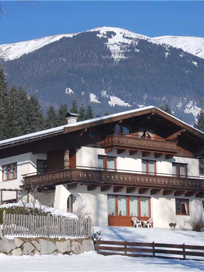 Ein traditionelles Chalet in einer verschneiten Landschaft mit Bergen im Hintergrund. Die Umgebung ist von Bäumen umgeben und strahlt eine ruhige Winteratmosphäre aus.