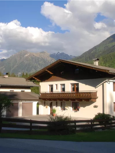 Ein charmantes Haus in den Bergen mit einem klaren Himmel und beeindruckenden Bergen im Hintergrund. Die Umgebung ist grün und einladend, ideal für Naturfreunde.