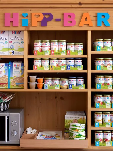 Eine Regale mit verschiedenen Babyprodukten der Marke HiPP. Dazu gehören Babynahrung, Brei und Snacks in einer gemütlichen Umgebung.