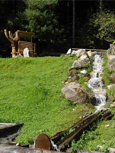 Ein kleiner Wasserfall fließt über Felsen in eine grüne Wiese. Im Hintergrund steht eine Holzstruktur, umgeben von Bäumen.