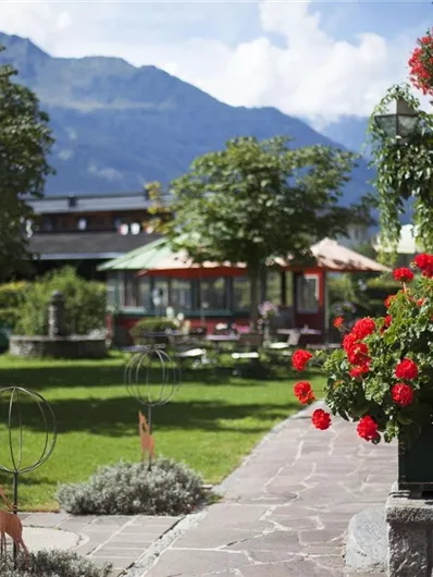 Ein schöner Garten mit blühenden roten Blumen und gepflegtem Rasen. Im Hintergrund sind Bäume und Berge sichtbar.