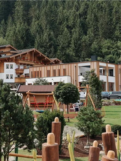 Ein modernes Gebäude aus Holz, umgeben von Bäumen und einer grünen Wiese. Im Vordergrund befindet sich ein Spielplatz mit verschiedenen Spielgeräten.