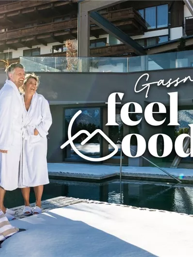 A couple in bathrobes stands relaxed by the pool of a hotel in the snow. The sun is shining, and the caption "Gassner feel good" is visible.