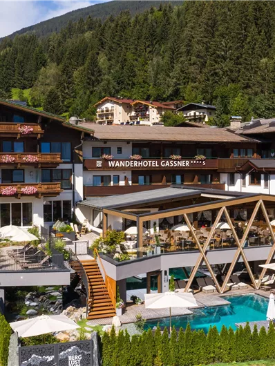 Ein modernes Hotel mit mehreren Etagen, umgeben von Bergen und Bäumen. Der großzügige Außenbereich umfasst einen Pool und gemütliche Terrassen.