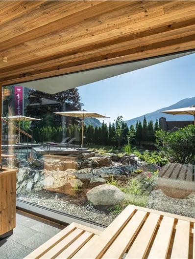 Ein moderner Wellnessbereich mit großen Fenstern und Blick auf die Berge. Draußen sind Liegestühle und eine gepflegte Gartenlandschaft zu sehen.