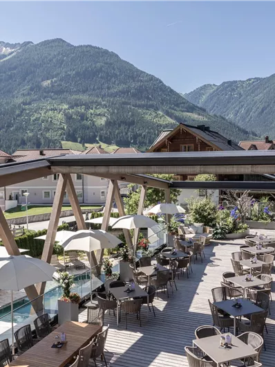 Een terras met moderne tafels en stoelen, omringd door bergen. De zonnige sfeer nodigt uit tot ontspannen.