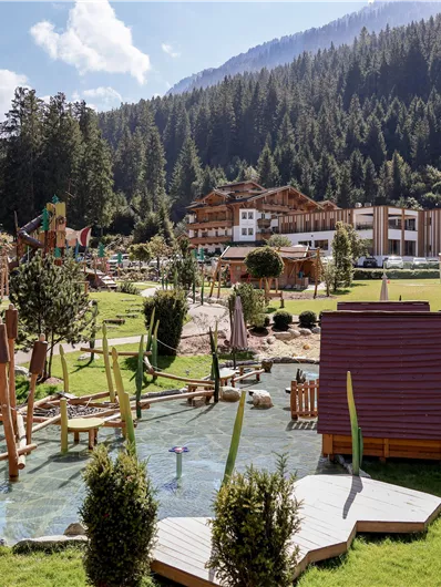 Ein liebevoll gestalteter Spielplatz mit Wasseranlagen und Spielgeräten. Im Hintergrund sind Wälder und Berge zu sehen, sowie ein gemütliches Gebäude.