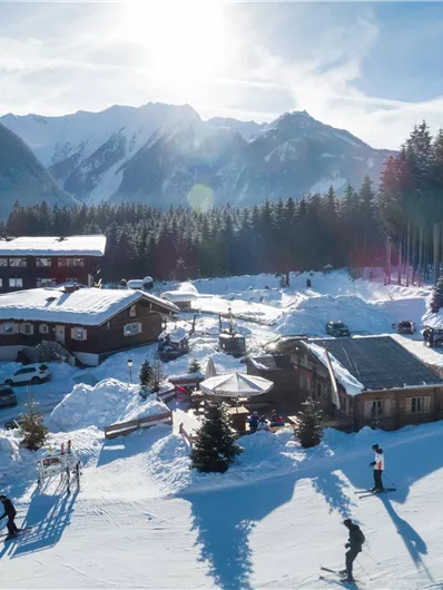 Eine malerische Winterlandschaft mit schneebedeckten Bergen und einem charmanten Gebäude. Im Vordergrund sind Skifahrer und schneebedeckte Bäume zu sehen.