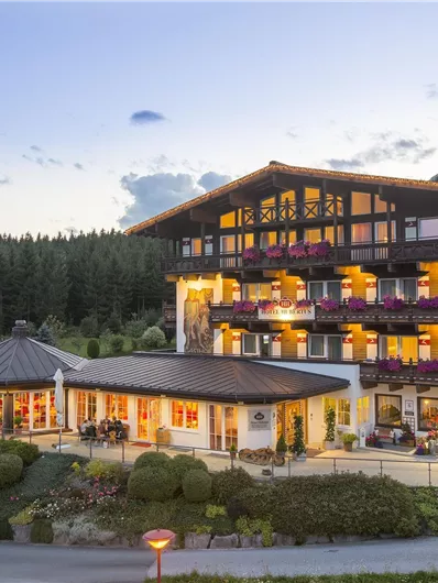 Ein stilvolles Hotel mit mehreren Etagen und Balkonen, umgeben von Bäumen und Bergen. Die Fassade ist hell beleuchtet und lädt zum Verweilen ein.