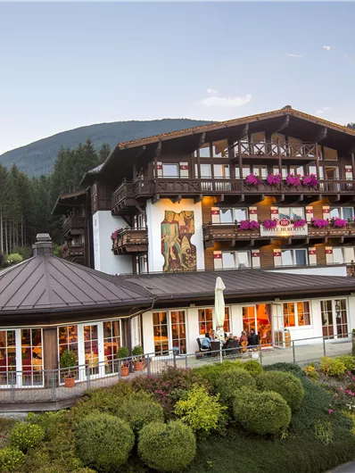 Ein traditionelles Alpenhotel umgeben von grünen Gärten und Bergen. Die Balkone sind mit blühenden Pflanzen dekoriert, und die Atmosphäre ist einladend.