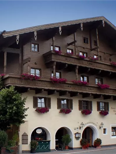 Een traditioneel houten huis met meerdere balkons en kleurrijke bloemen. Op de achtergrond zijn zachte heuvels en bomen te zien.