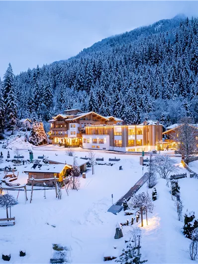 Ein malerisches Berghotel, umgeben von schneebedeckten Bäumen und Bergen. Die Gebäude sind warm erleuchtet und schaffen eine einladende Atmosphäre im Winter.