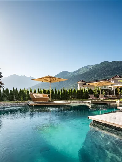 Ein schöner Pool mit klarem Wasser, umgeben von grünen Pflanzen und Sonnenschirmen. Im Hintergrund sind sanfte Berge und ein blauer Himmel zu sehen.