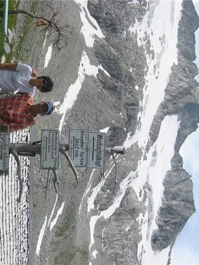 Twee personen staan voor een indrukwekkend berglandschap met besneeuwde toppen. Op de voorgrond zijn borden zichtbaar die informatie verstrekken.