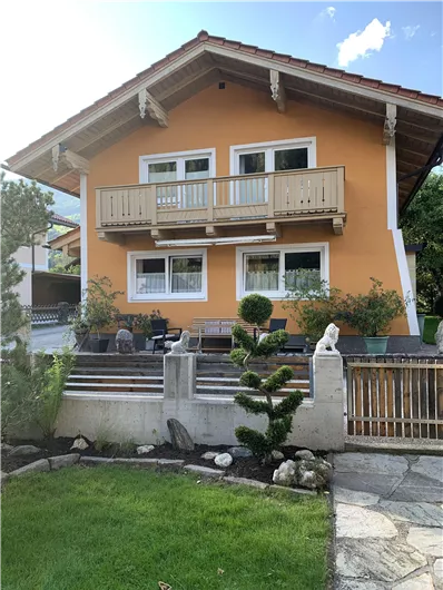A beautiful orange house with a balcony and large windows. The garden is well-maintained with a small rock garden and green plants.