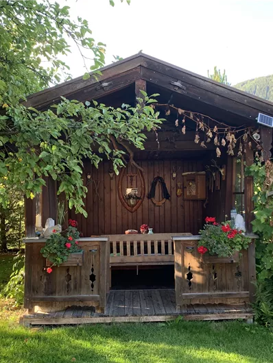 A cozy garden shed surrounded by green plants and flowers. Next to the shed is a grill.